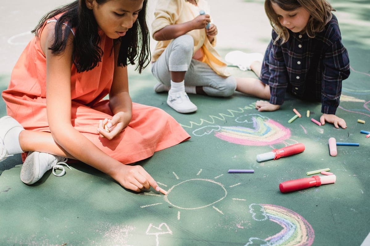 Three children drawing rainbows and sunshine in colored chalk on pavement