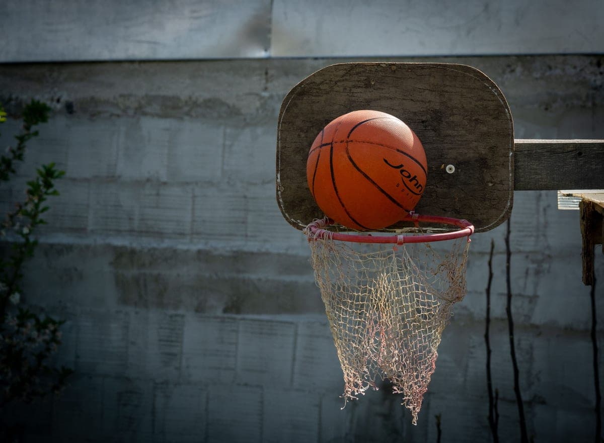 A basketball resting on an outdoor rim with a worn net and a wooden backboard