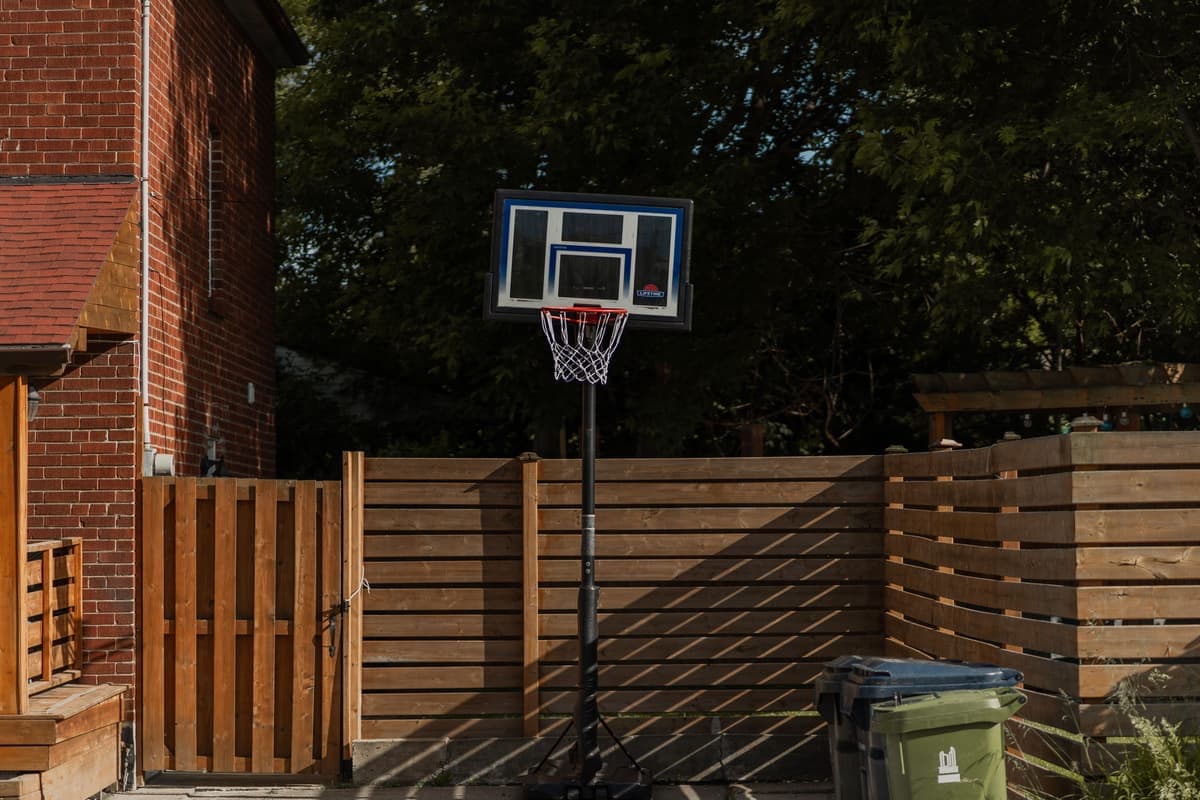 A backyard basketball hoop on a pole beside a brick house and a wooden fence